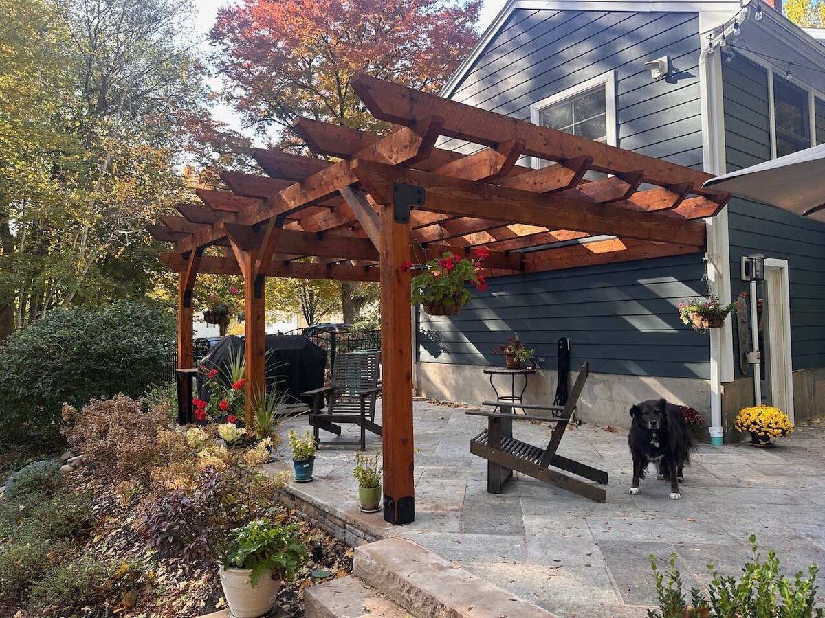 Wooden pergola over stone patio with seating and hanging planters in Ridgewood, NJ by Fairmount Heights Homes.